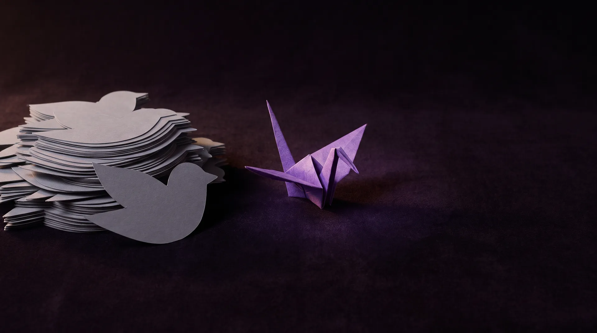 Kasa samanlaisia harmaita paperileikkeitä ja erillään yksi violetti origamilintu